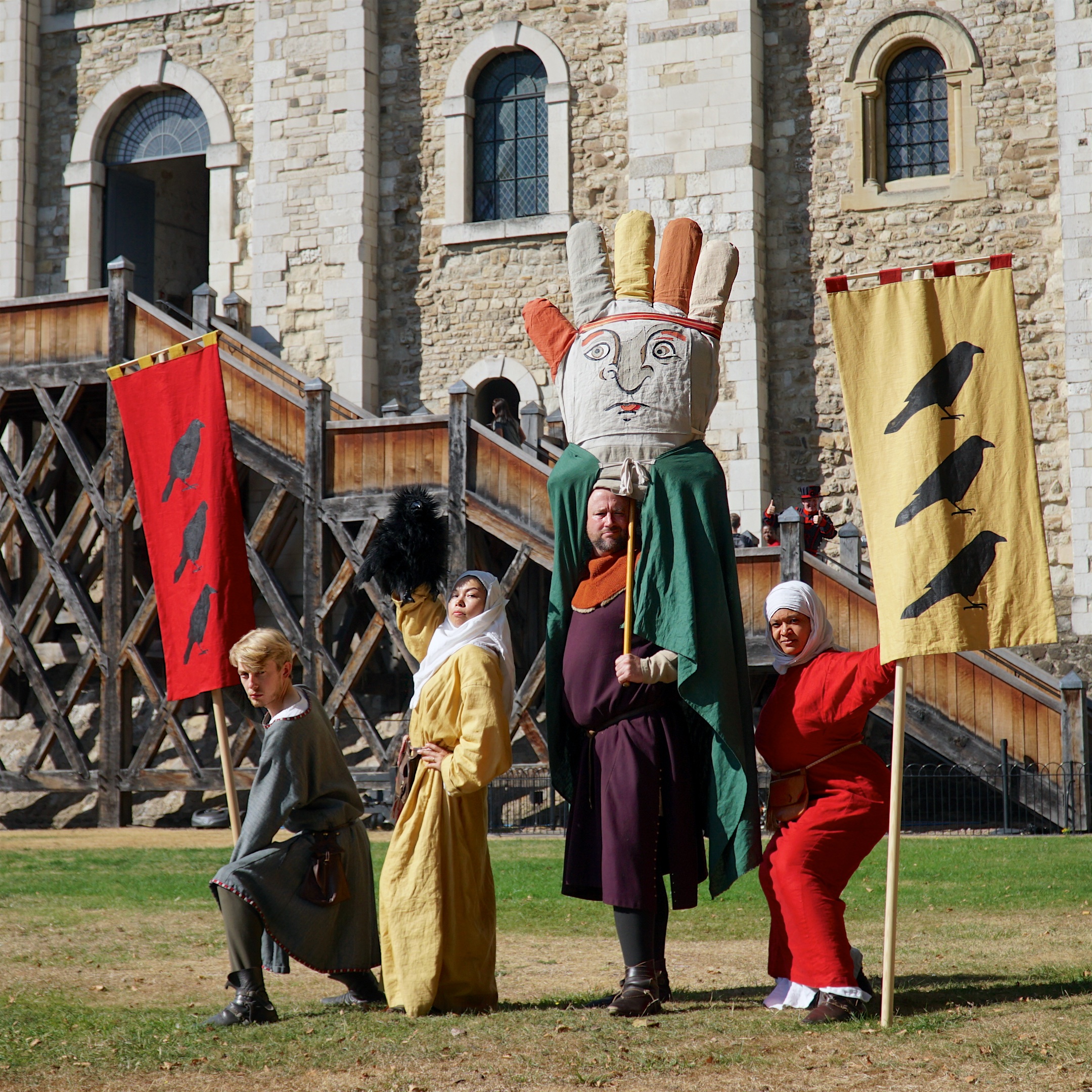 Four medival characters with flags and a giant glove puppet stand in front of the White Tower at the Tower of London in the Sun